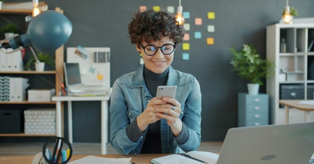 Mulher sorri enquanto mexe no celular em um escritório criativo de comunicação, com post-its coloridos ao fundo